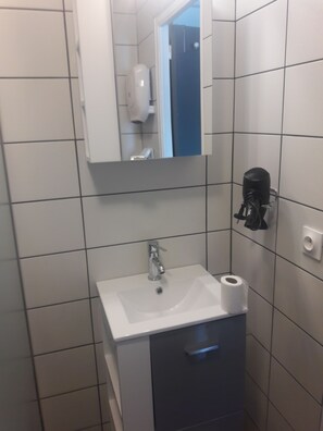 Une salle de bain avec un lavabo blanc, un miroir et un sèche-cheveux noir fixé au mur.