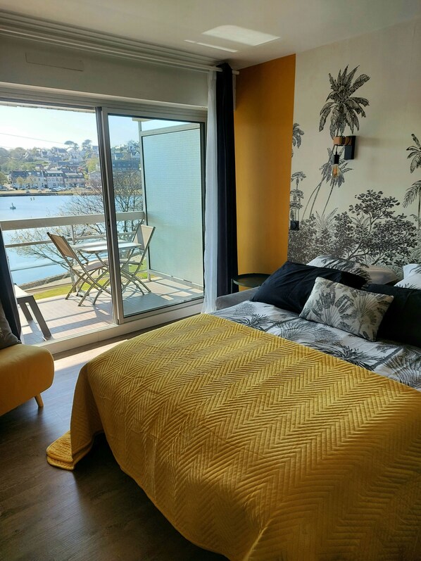 Une chambre dotée d’un grand lit, donnant sur un balcon avec des chaises, et dont un des murs est décoré d’une fresque représentant des plantes tropicales.