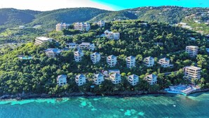 An aerial view of a coastal resort with multiple buildings, lush greenery, and clear turquoise waters.