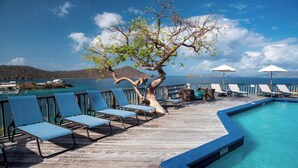 A wooden deck with lounge chairs and a pool, overlooking a body of water with boats and a mountainous backdrop.