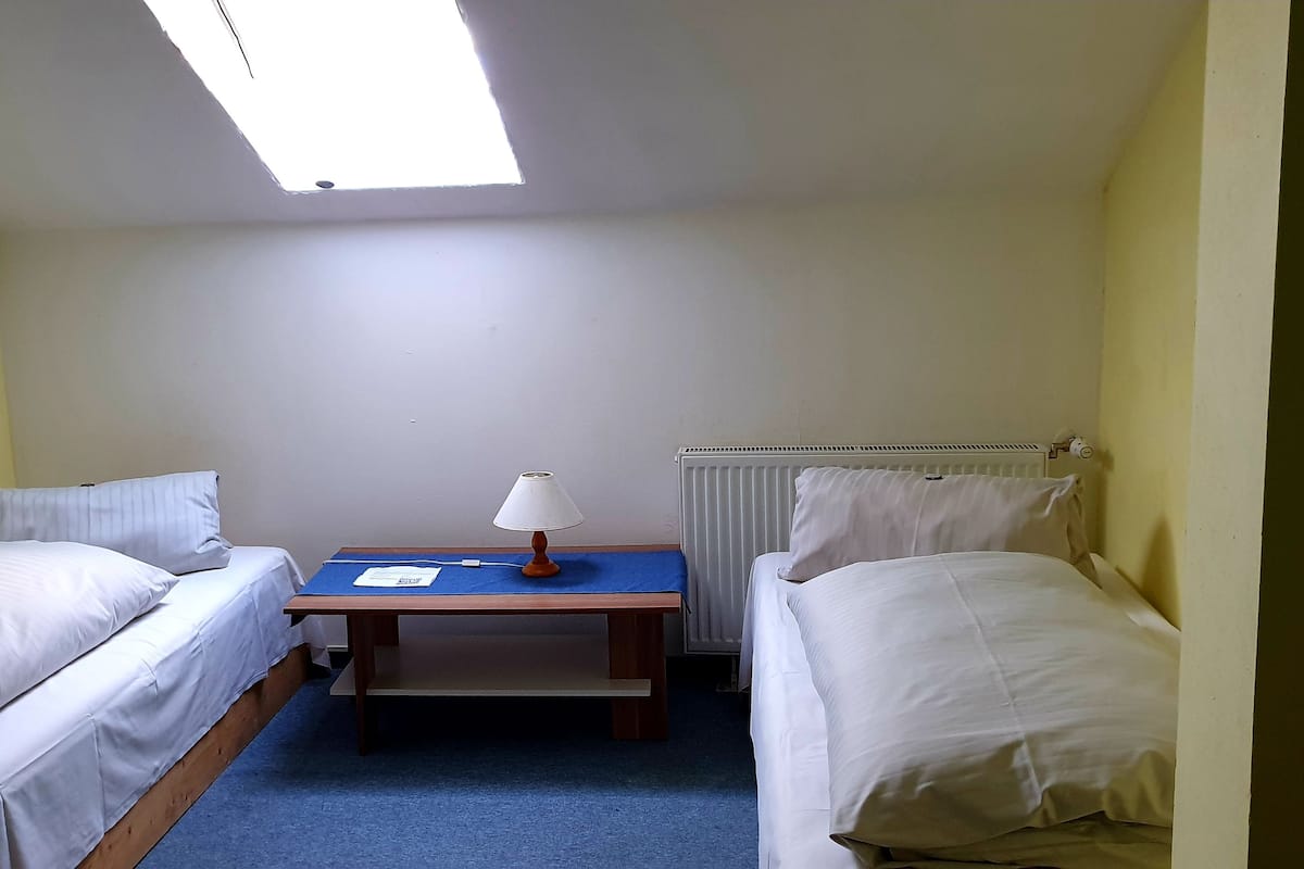 A small, single-room hotel room with two single beds, a small table with a lamp, and a radiator.