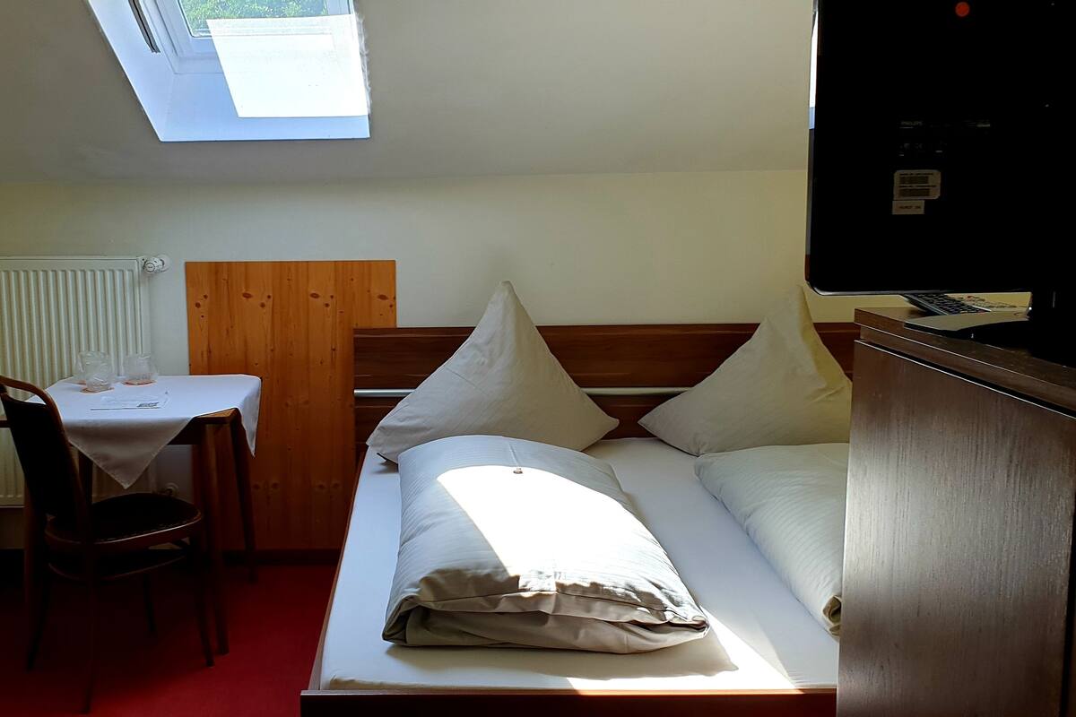 A small, well-lit room with a bed, a desk, and a chair. There is a skylight above the desk, a radiator on the wall, and a wooden headboard.