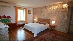 Een slaapkamer met een stenen muur, een houten bed, twee nachtkastjes met lampen, een raam met gordijnen en een houten dressoir met een vaas rode rozen.