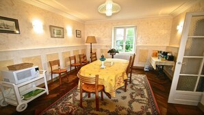 Een eetkamer met een ronde tafel, stoelen, een kroonluchter, een raam en een zichtbare keuken door een open deur.