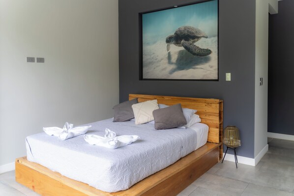 A neatly made bed with a wooden headboard, a framed picture of a sea turtle on the wall, and a small wicker chair beside the bed. - Lilan Nature, Modern House N°1 with private swimming pool, Tennis and Pickleball (Cahuita)