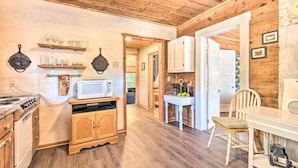 A wooden interior with a kitchen area featuring a stove, microwave, and cabinets. There is a dining table with chairs and a small white cabinet with a blender. - Updated Cabin w/ Porch, Mins to Cossatot River (Wickes)