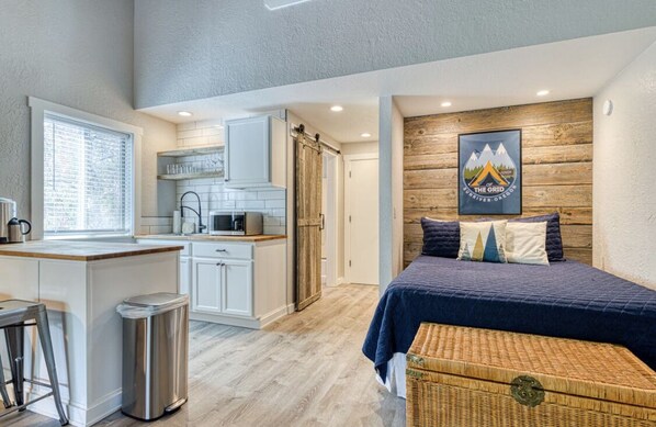 A compact room with a kitchenette, a bed with a headboard, and a wooden accent wall.