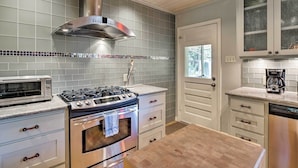 Een keuken met een gasfornuis, oven, magnetron en een roestvrijstalen afzuigkap. Er zijn witte kasten met glazen deuren en een houten aanrecht.