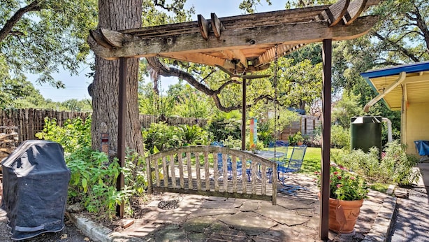 Een houten pergola met een bank, omgeven door een stenen terras en weelderige beplanting.