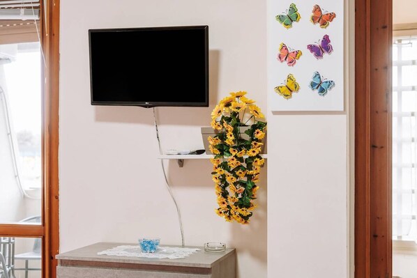 A room with a flat-screen TV mounted on the wall, a shelf with decorative items, and a table with a bowl and a glass.