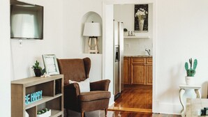 Ein gemütliches Wohnzimmer mit einem braunen Sessel, einem Holzregal mit dem Schriftzug 'BE HAPPY' und einer durch einen offenen Durchgang einsehbaren Küche.