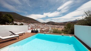 Una piscina con muelle de madera y sillones de descanso, rodeada por un paisaje montañoso y un pueblo a lo lejos.