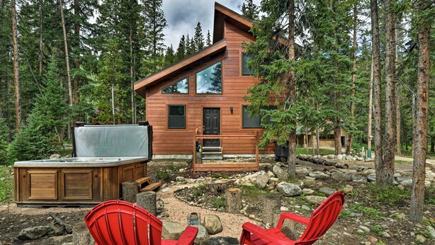 Una cabaña de troncos moderna con jacuzzi, dos sillas Adirondack rojas y una terraza con vistas a un área boscosa.