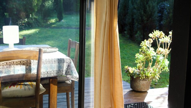 A wooden deck with a table and chairs, a potted plant, and a view of a grassy area through a window with curtains. - Studio, garden, jacuzzi, sauna, yoga, massages (Soorts-Hossegor)