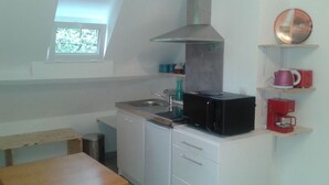 A small kitchen with a sink, a stove, and a microwave. There is a window above the sink and a shelf with a kettle and cups. - ----- Green Studio ----- (Curemonte)