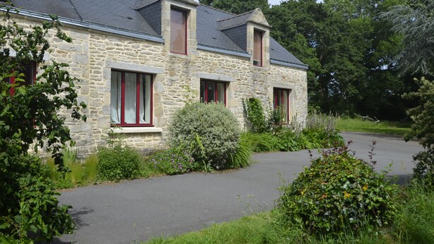 Ein Haus aus Stein mit einem geneigten Dach, mehreren Fenstern mit roten Rahmen und einer gepflasterten Auffahrt.
