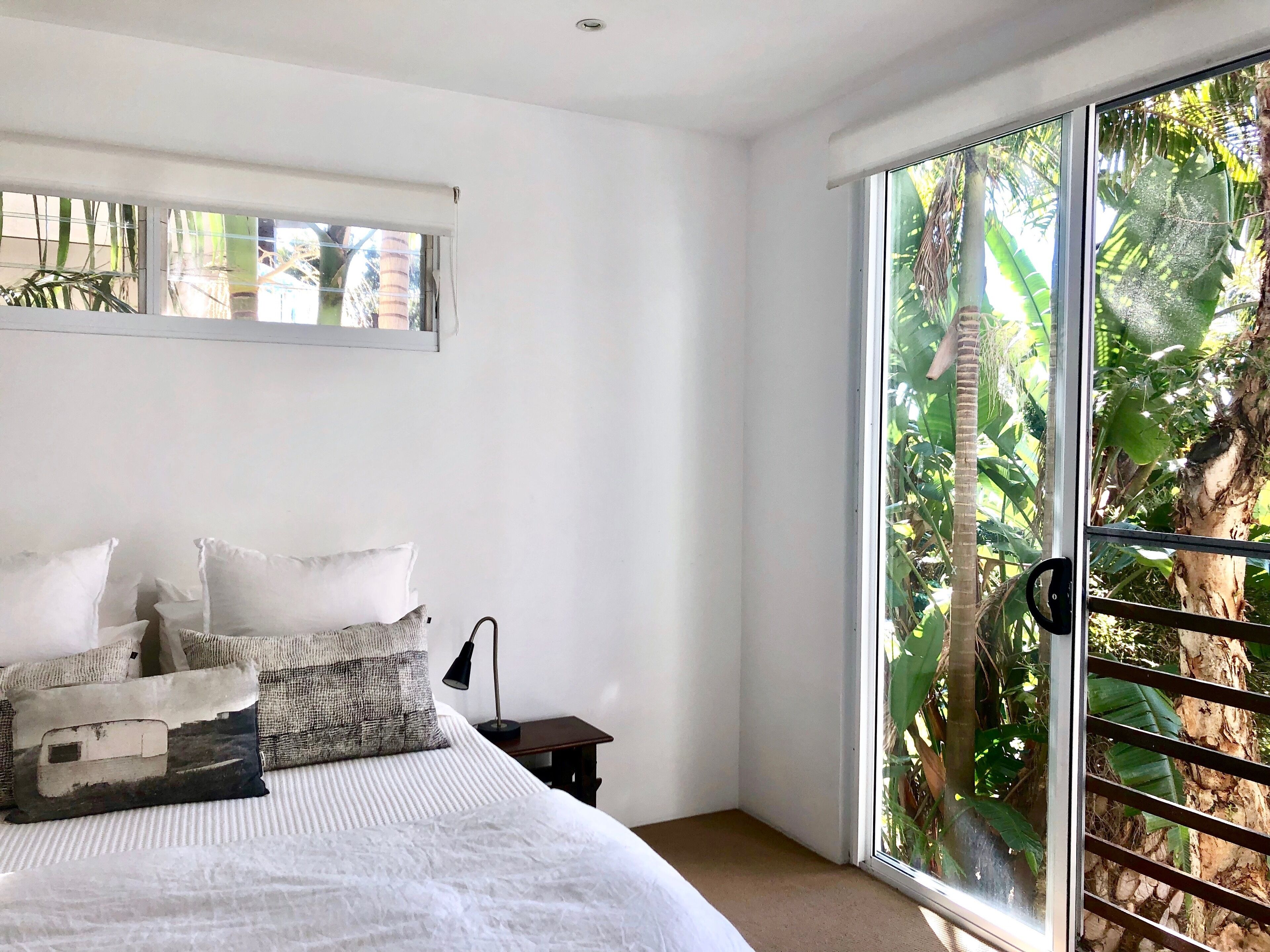 A bedroom with a large bed, a nightstand, a lamp, and a sliding glass door leading to a tropical garden.