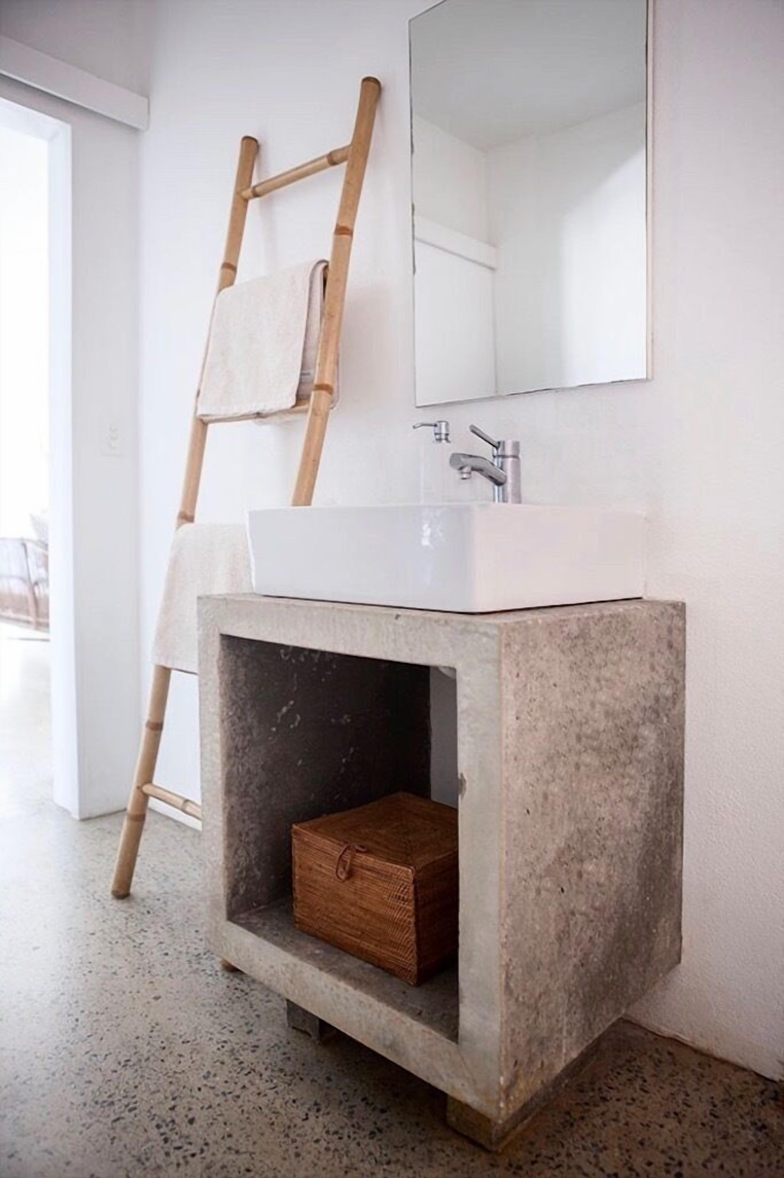Ein Badezimmer mit einem weißen Waschbecken, einem Spiegel, einer Holzleiter und einer kleinen Holzschatulle in einem Betonschrank.