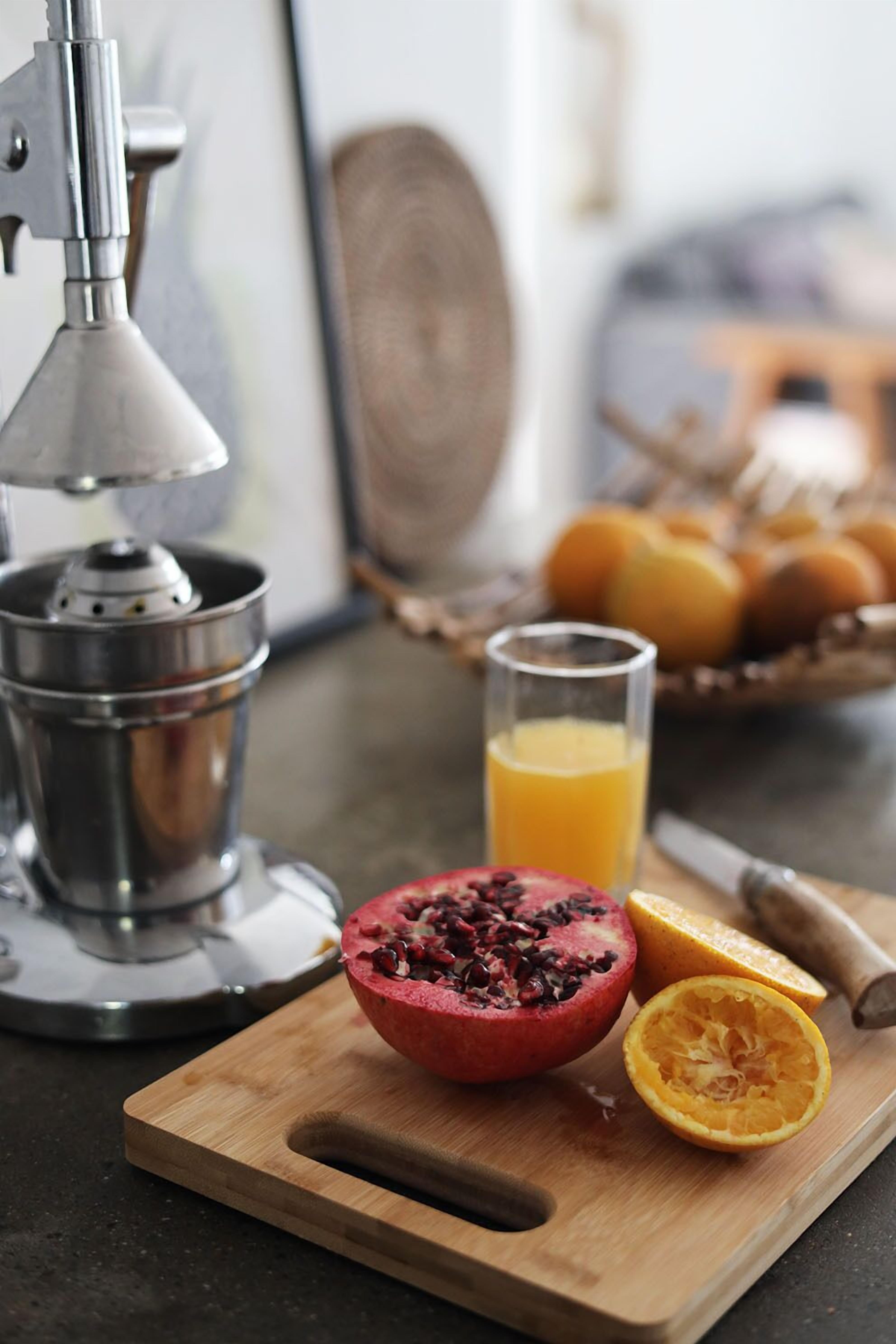 A kitchen counter with a juicer, a pomegranate cut in half, an orange half, and a glass of orange juice.
