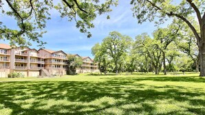 A courtyard with a grassy lawn, trees, and a multi-story building with balconies. - Waterwheel Retreat | Pet Friendly | 3rd Floor (New Braunfels)