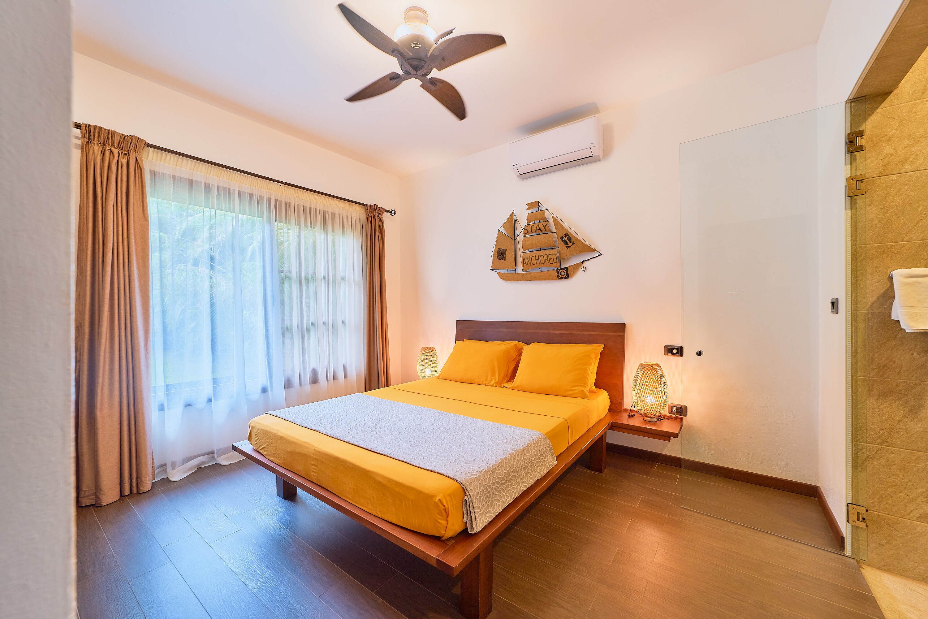 A bedroom with a bed, curtains, a ceiling fan, an air conditioning unit, a wall-mounted shelf with a decorative item, and a wooden floor.