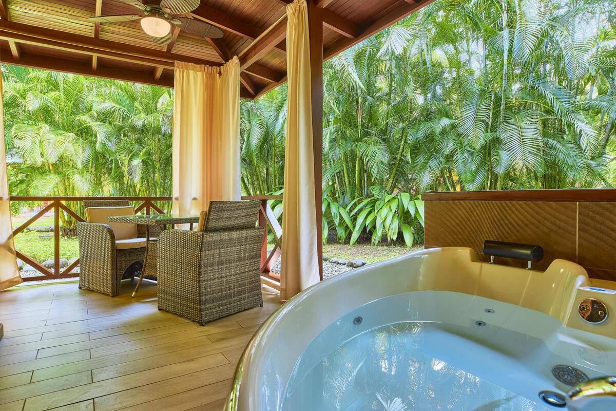 A covered outdoor area with a hot tub, wicker furniture, and lush tropical vegetation.