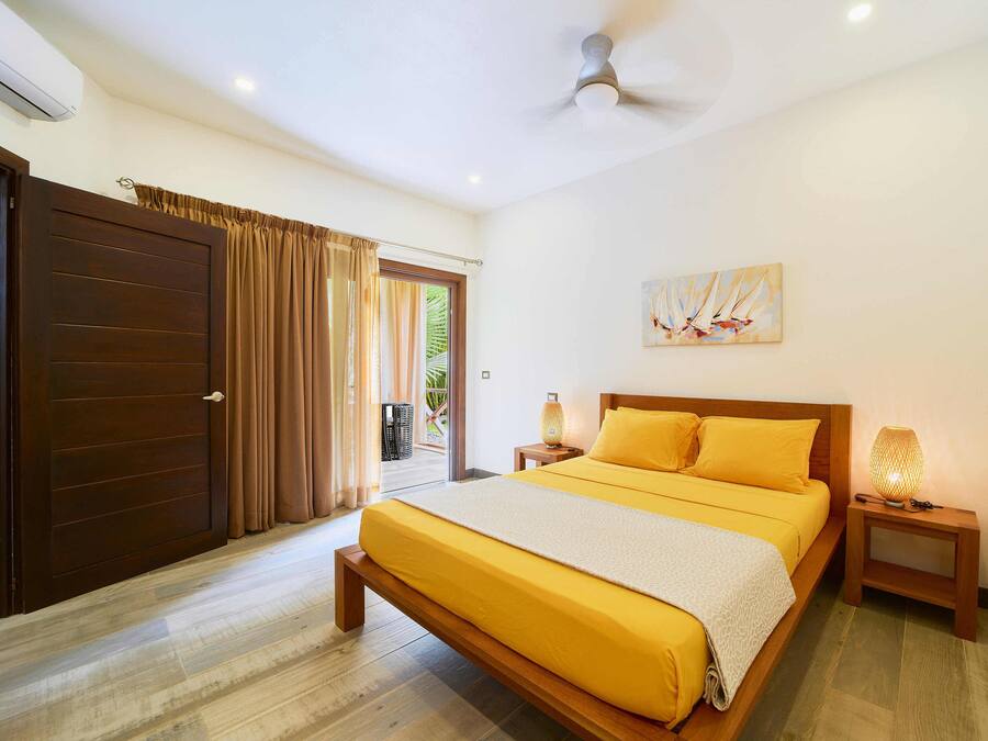 A bedroom with a large bed, wooden headboard, a ceiling fan, a nightstand with a lamp, and a door with curtains.