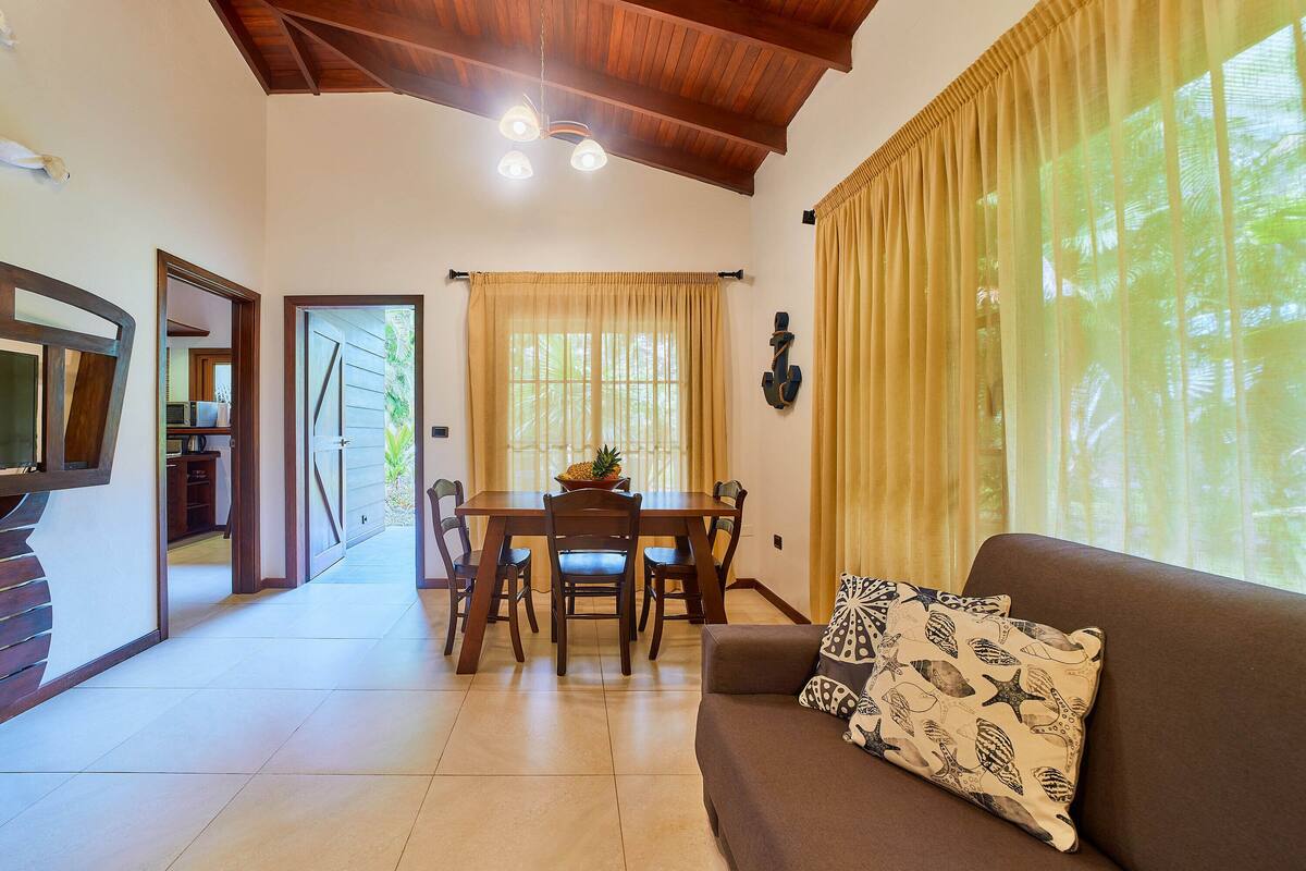 A spacious living area with a wooden ceiling, a dining table with chairs, a sofa with patterned cushions, and a large window with curtains.