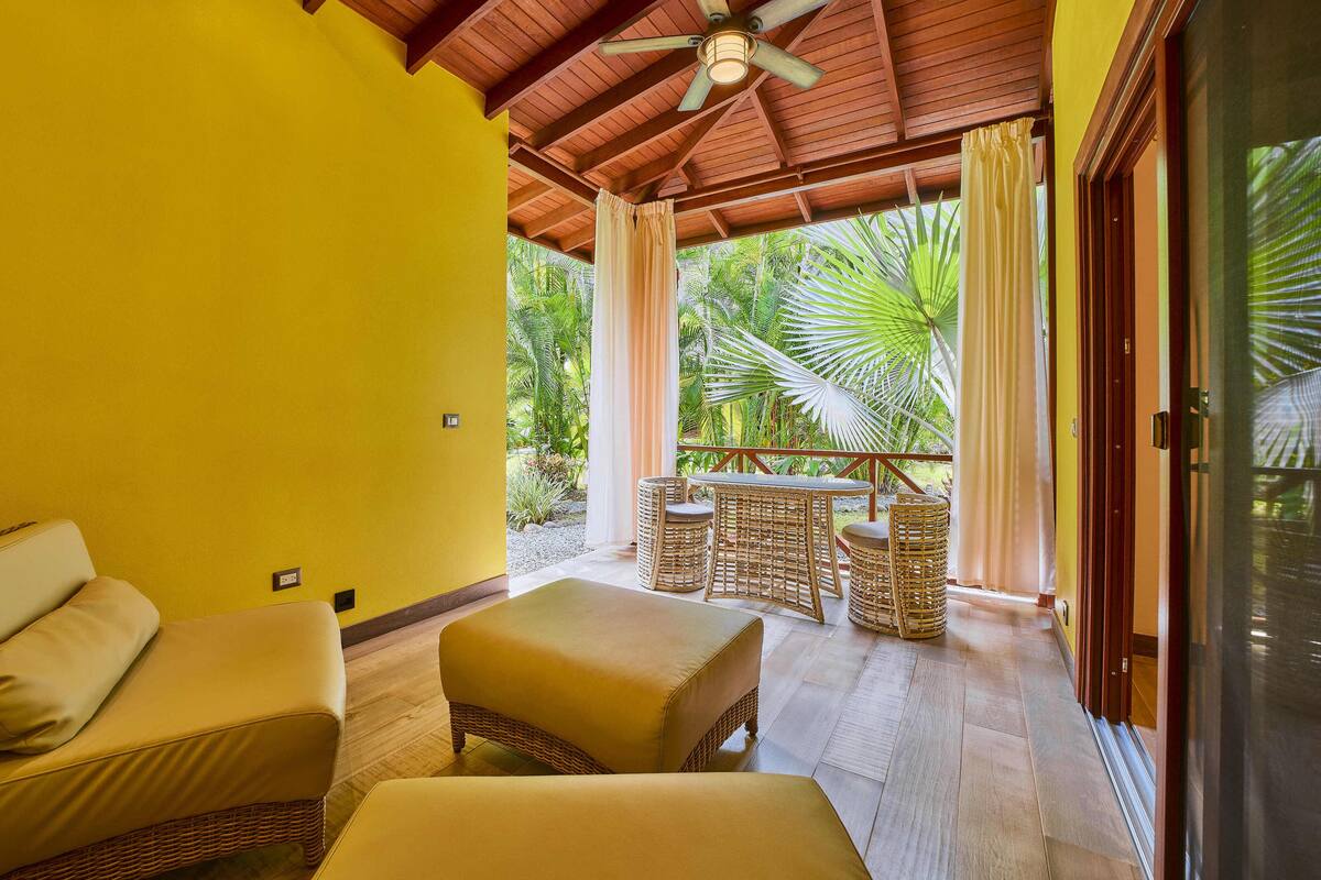 A room with a ceiling fan, wicker furniture, and a view of tropical foliage.