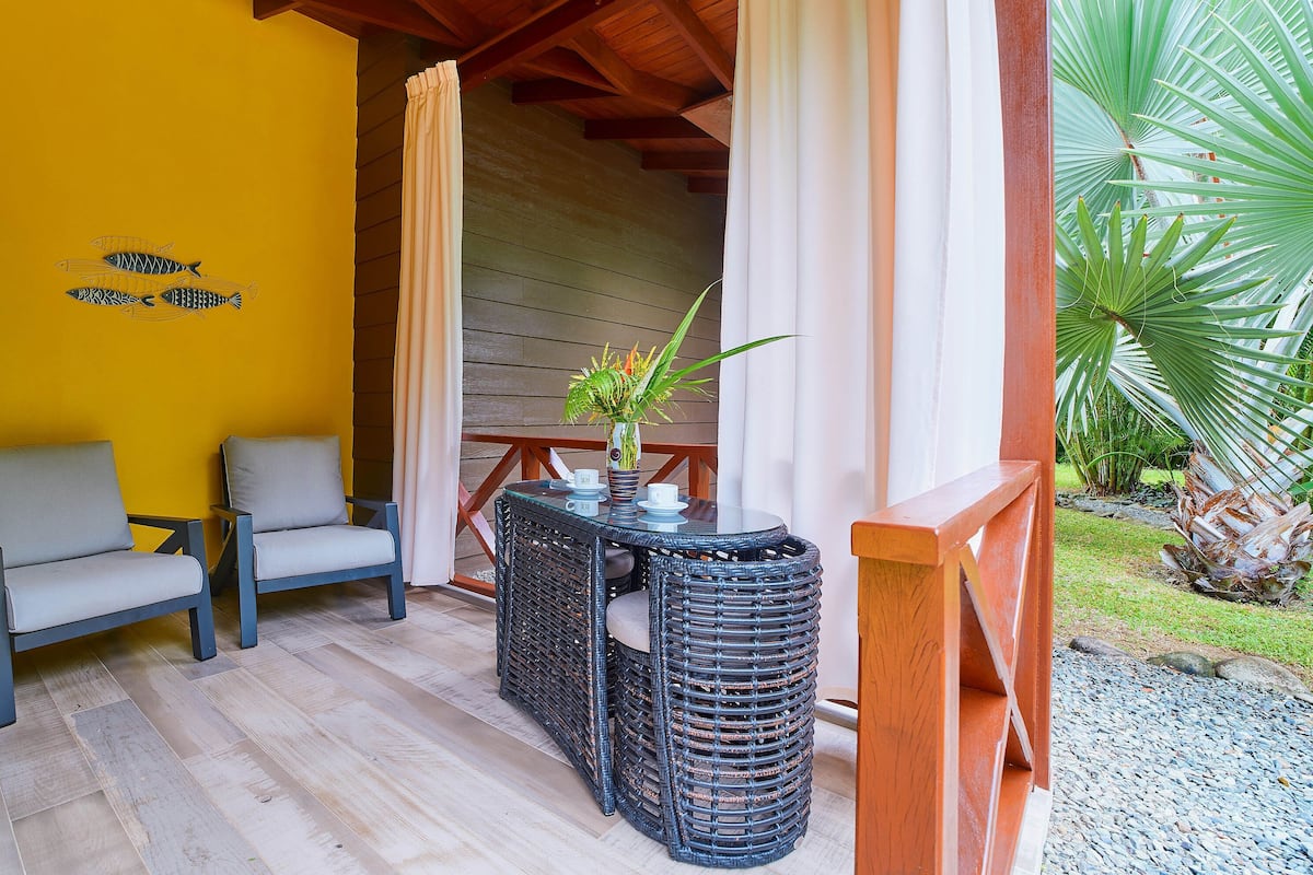 A covered outdoor seating area with wicker furniture and a table set for two, featuring a vase with flowers.