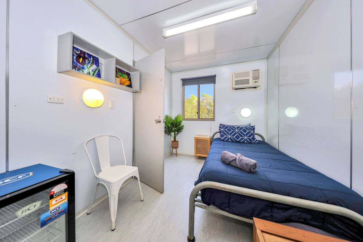 A compact room with a bed, a chair, a shelf with artwork, a window, and a radiator.