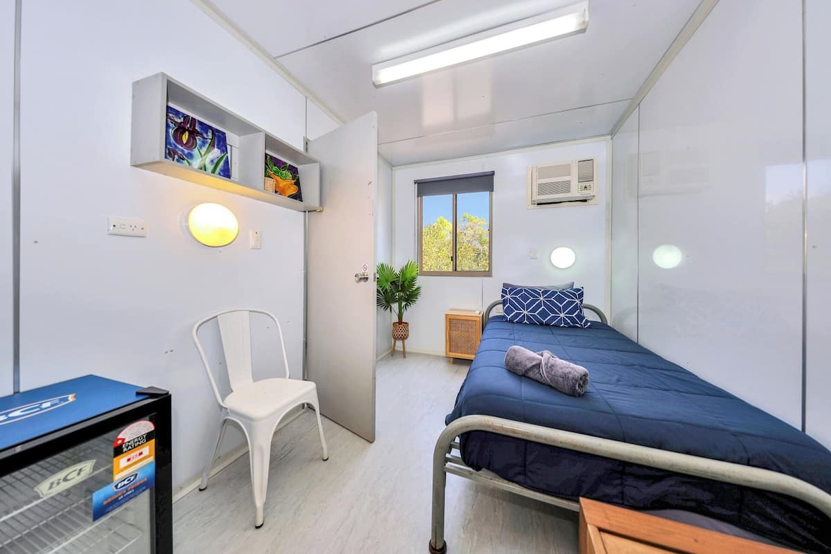 A compact room with a bed, a chair, a shelf with artwork, a window, and a radiator.