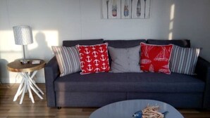 Een woonkamer met een grijze bank, rode en witte kussens met een patroon, een ronde salontafel met houten poten, een klein bijzettafeltje met een lamp, en een wand met maritieme decoratie.