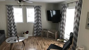 Een woonkamer met een houten vloer, een zwarte bank, een ronde salontafel, een zwarte fauteuil, een aan de muur bevestigde televisie en gordijnen met een chevronsmotief.