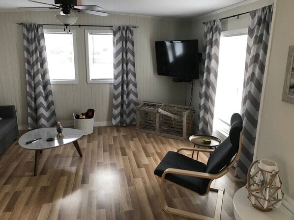A living room with a wooden floor, a black sofa, a round coffee table, a black armchair, a TV mounted on the wall, and chevron-patterned curtains.