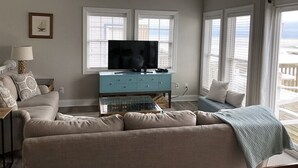 Ein Wohnzimmer mit Fernseher auf einem blauen Schrank, beigen Sofas und großen Fenstern mit Jalousien.