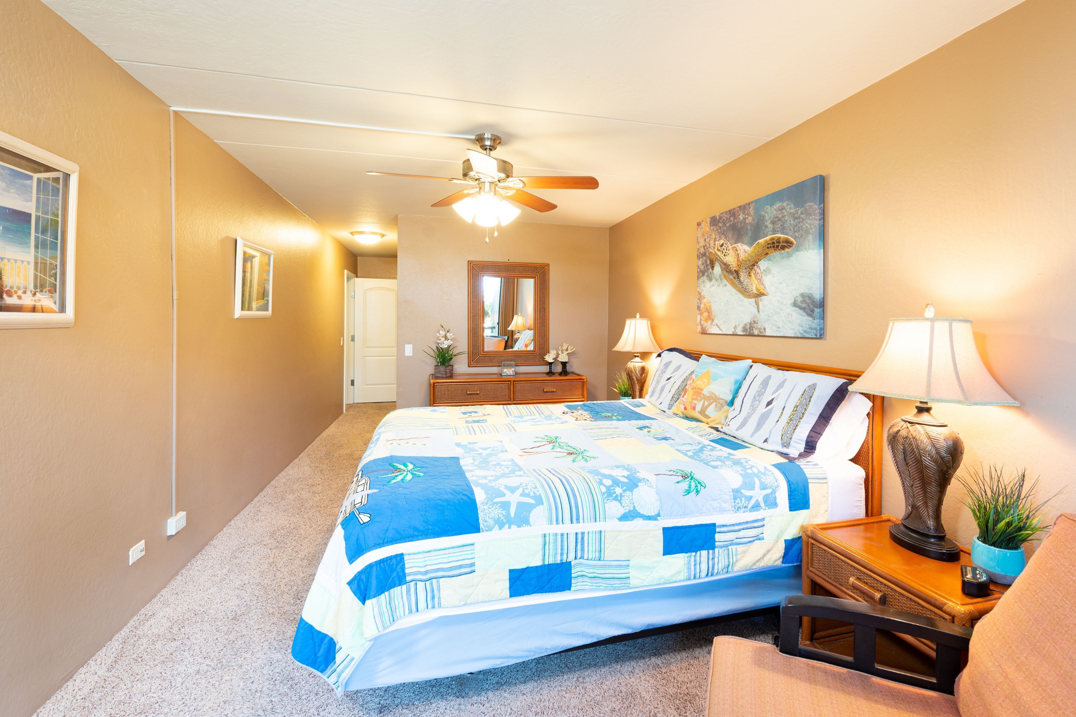 A bedroom with a large bed, a ceiling fan, a nightstand with a lamp, a mirror, and a painting of a sea turtle.