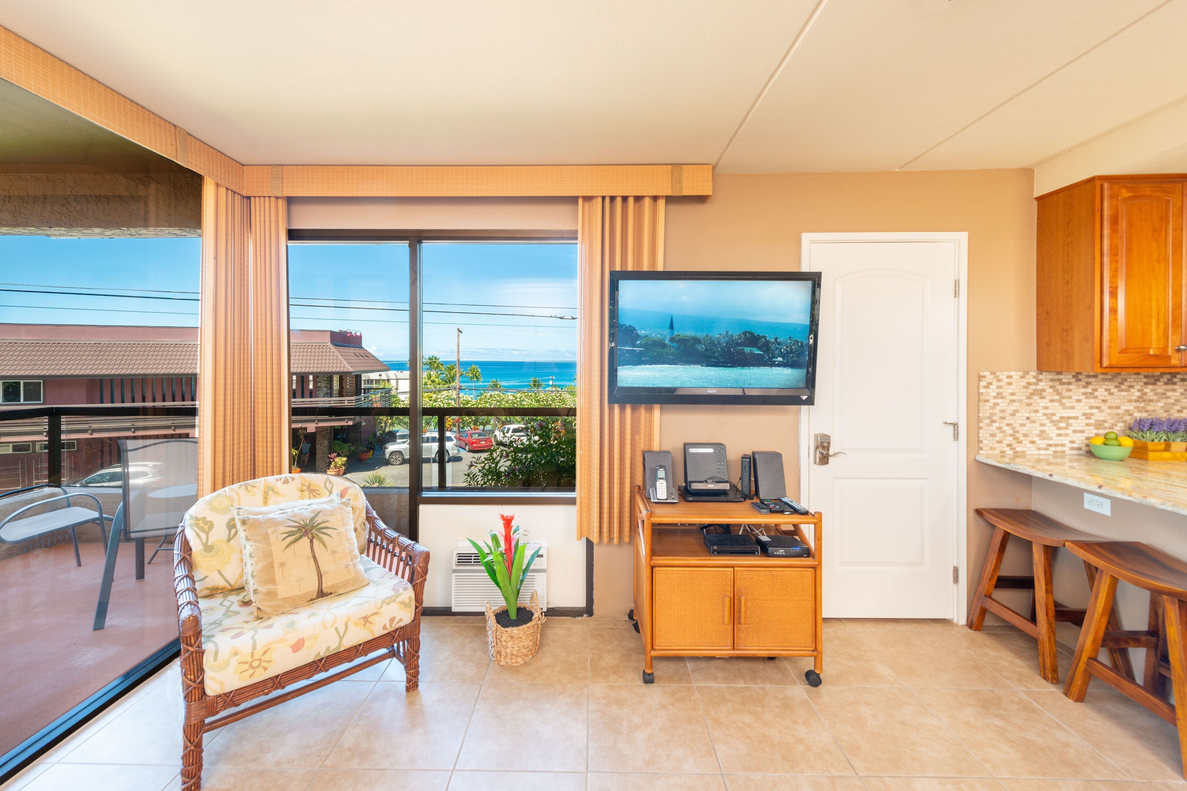 A modern living room with a large window, a flat-screen TV, a wicker sofa, a small wooden table, and a kitchen area with wooden cabinets.