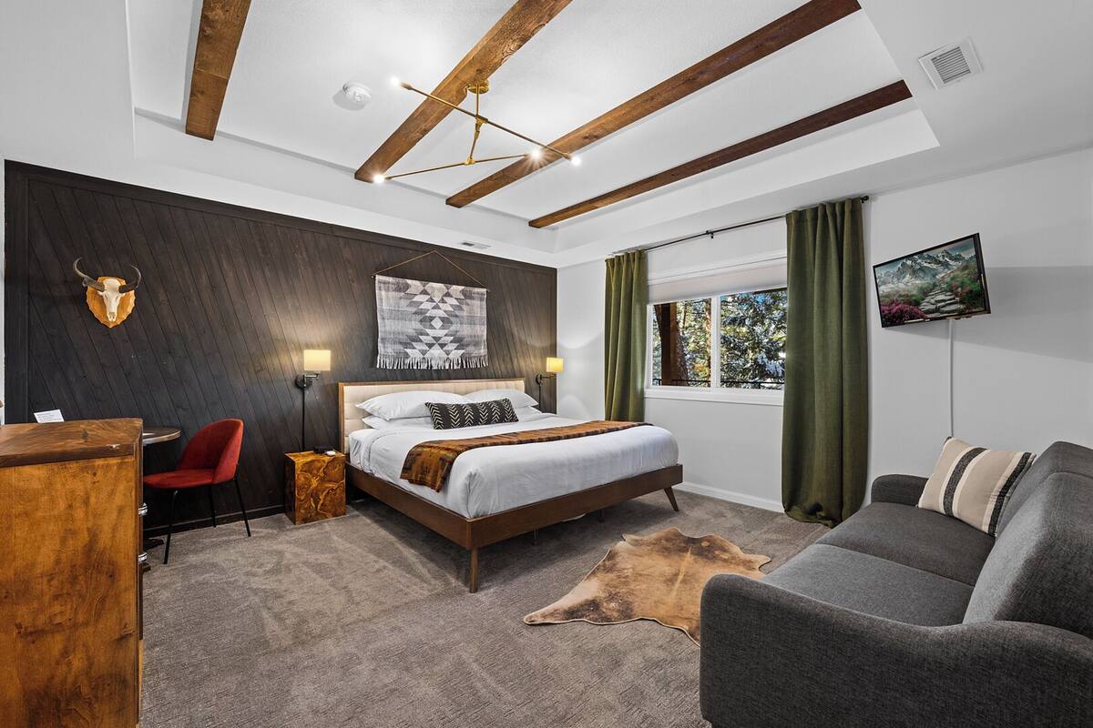 Quarto moderno com teto de madeira, cama com roupa de cama branca, escrivaninha com cadeira vermelha e uma janela grande com cortinas.