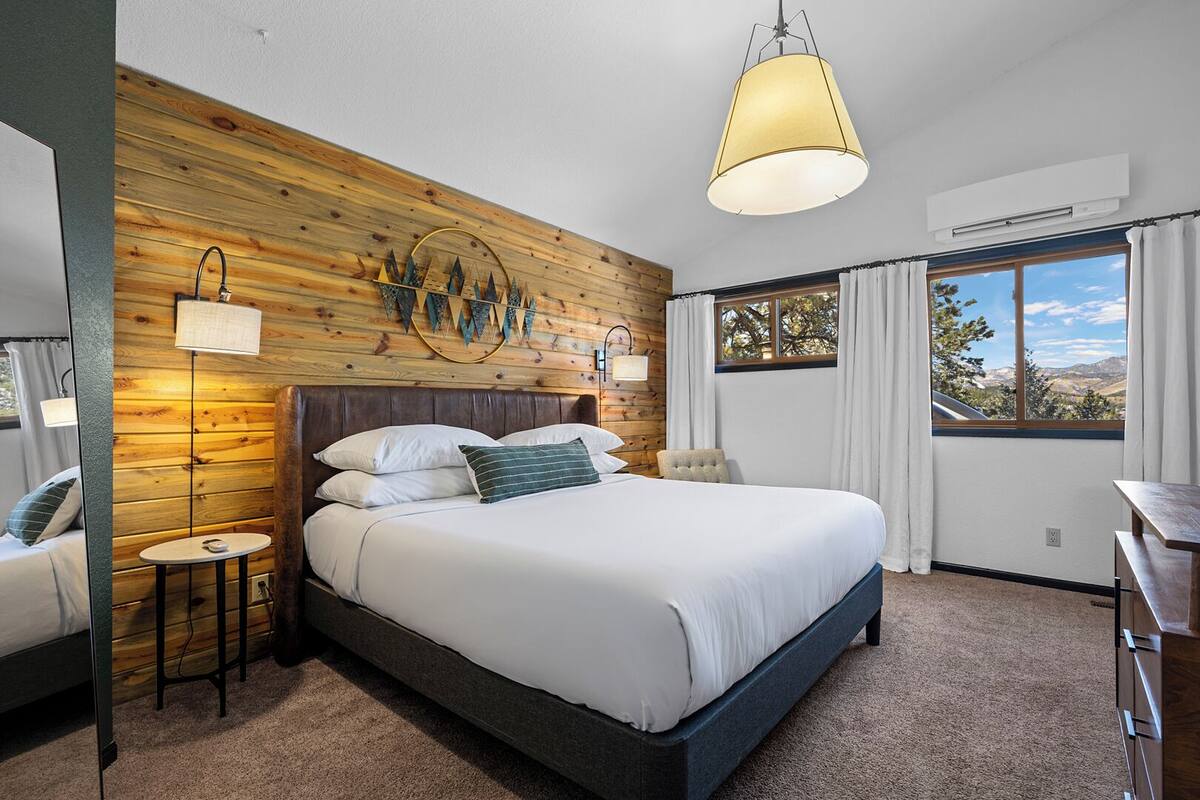 Quarto moderno com cabeceira de madeira, uma cama grande com roupa de cama branca, uma mesa redonda e vista para o exterior pela janela.