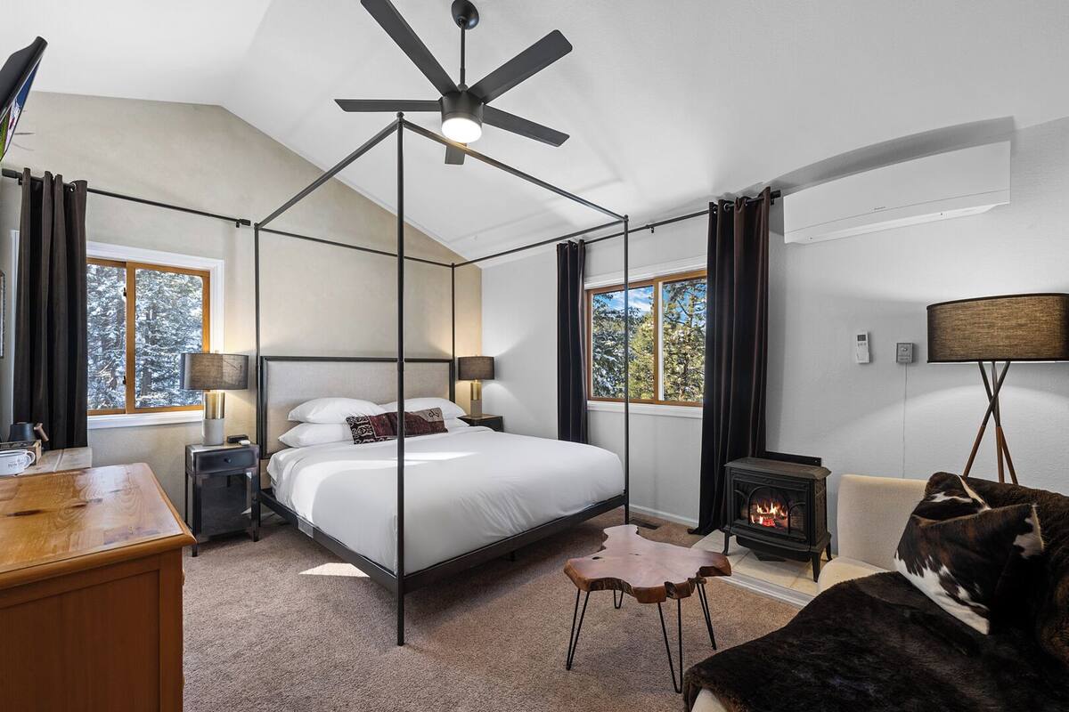 Quarto com cama de dossel, ventoinha no teto, lareira e vista para árvores pelas janelas.