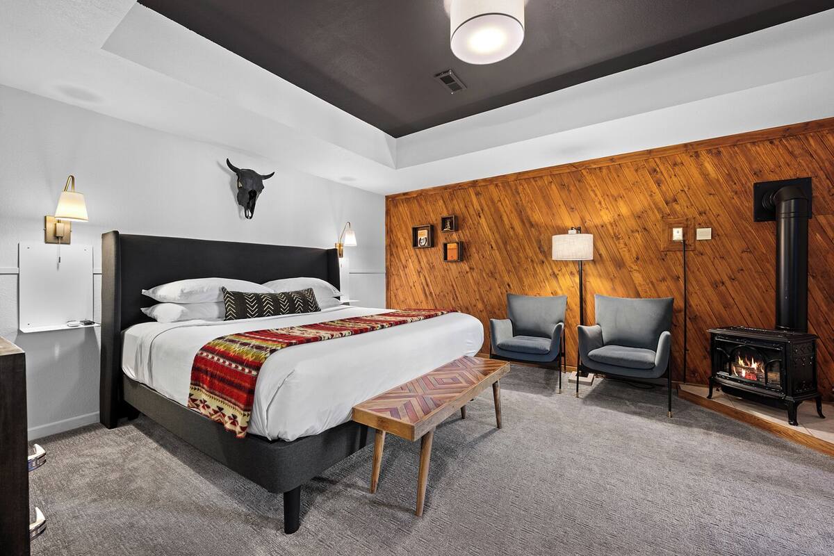 Quarto de hotel moderno com uma cama grande, duas poltronas, um painel de parede de madeira, teto preto e lareira a lenha.