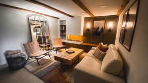 A cozy living room with a sofa, armchair, and coffee table. There is a fireplace with a shelf of books and decorative items. - Juniper Lodge & Treehouses (Evergreen)