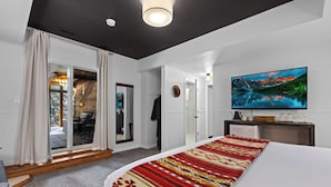 A modern hotel room with a large bed, a flat-screen TV, a sliding glass door to a balcony, and a view of a mountainous landscape. - Juniper Lodge & Treehouses (Evergreen)