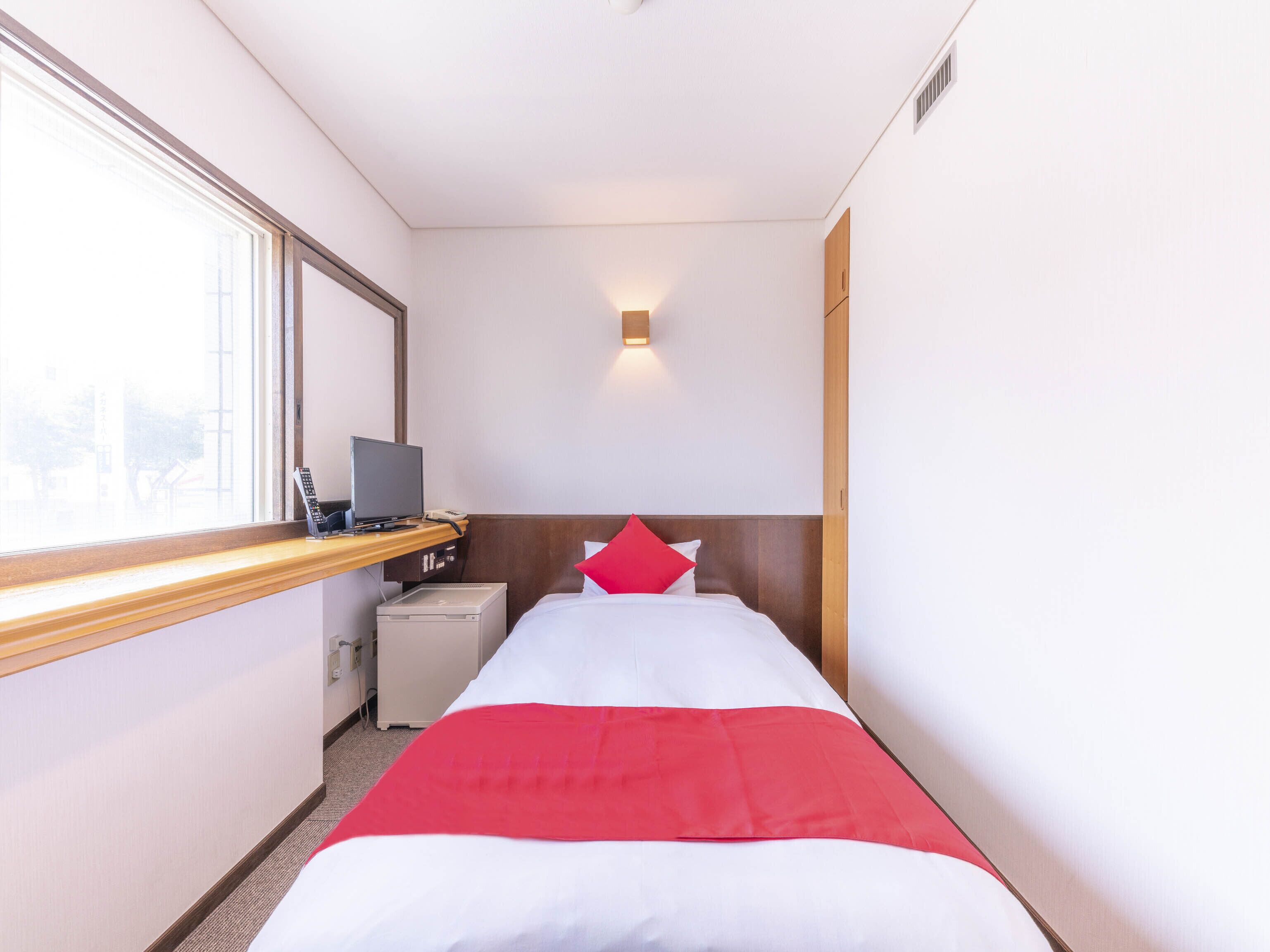 A hotel room with a bed, a desk with a computer, a small desk lamp, and a window.