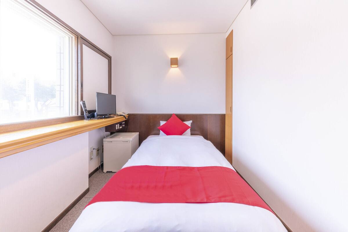 A hotel room with a bed, a desk with a computer, a small desk lamp, and a window.