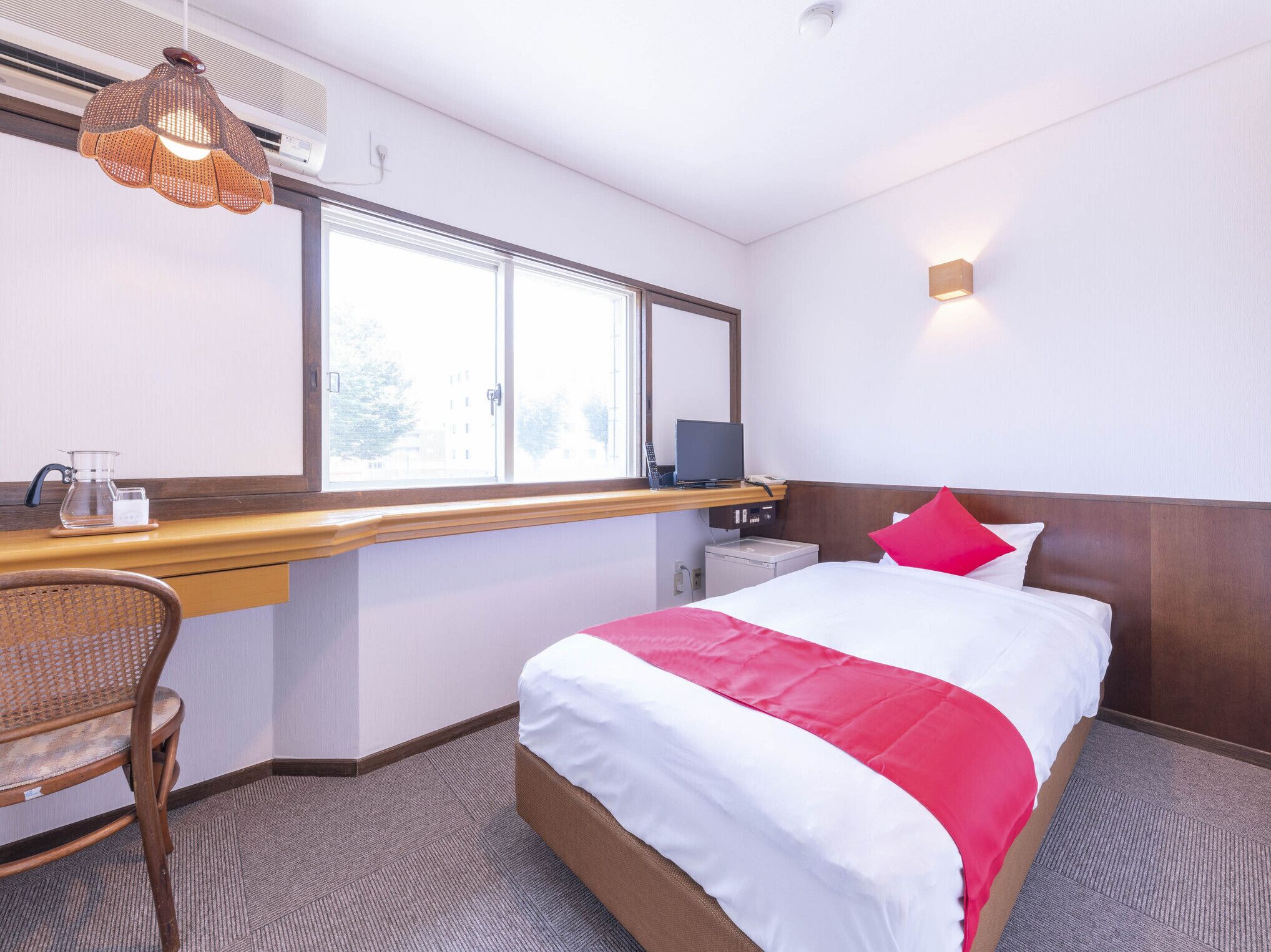 A hotel room with a bed, a desk, a chair, a window, and a wall-mounted light.