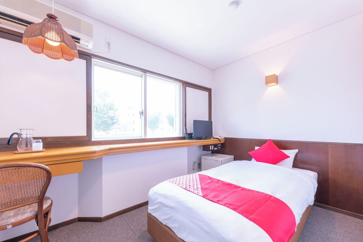 A hotel room with a bed, a desk, a chair, a window, and a wall-mounted light.