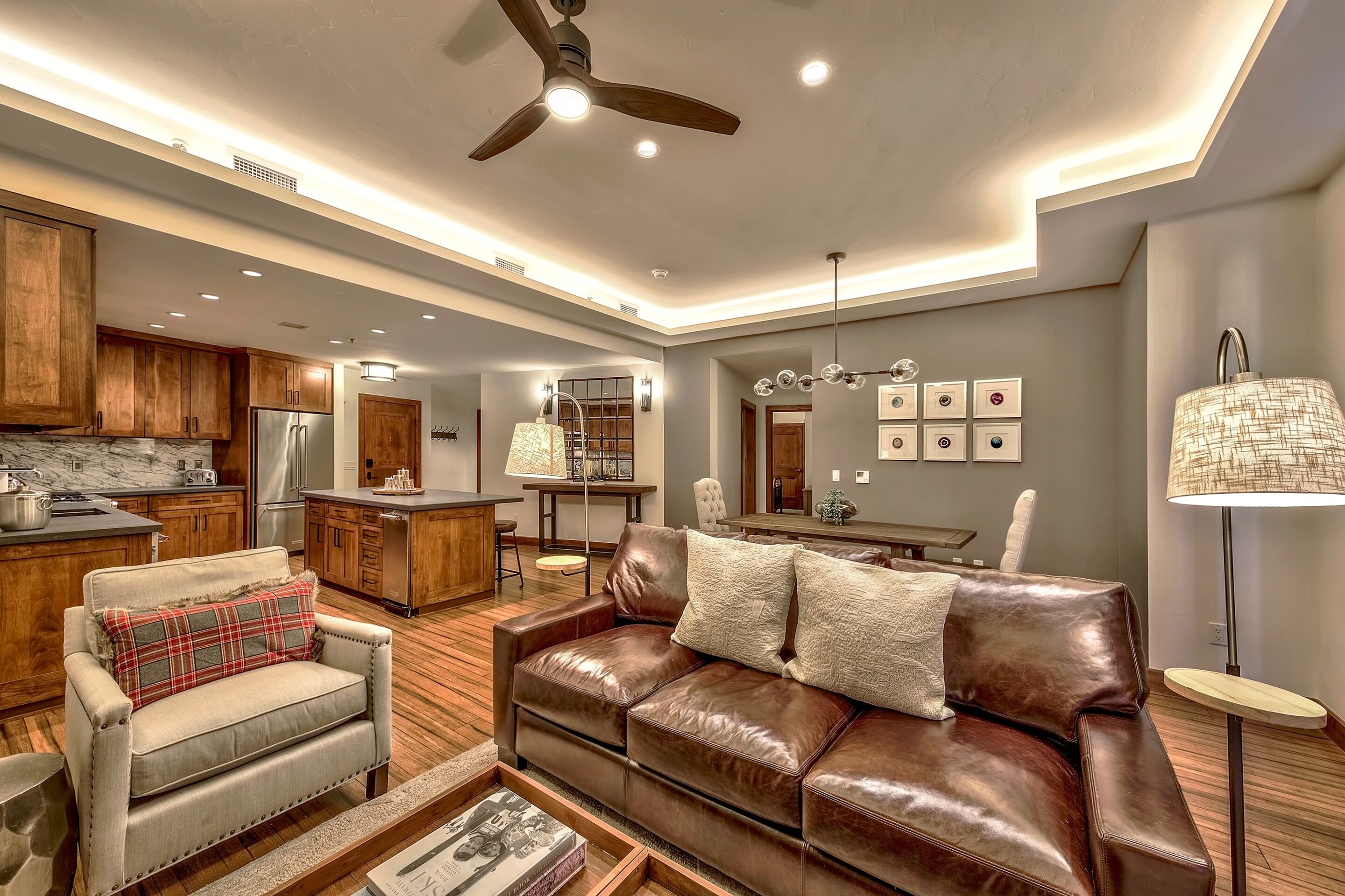 A modern living room with a leather sofa, a wooden dining table, and a kitchen area with wooden cabinets and a refrigerator.