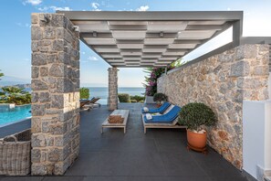 Un espace de détente extérieur, revêtu de pierre, avec une piscine, deux coins salons dotés de coussins bleus et une pergola au-dessus.